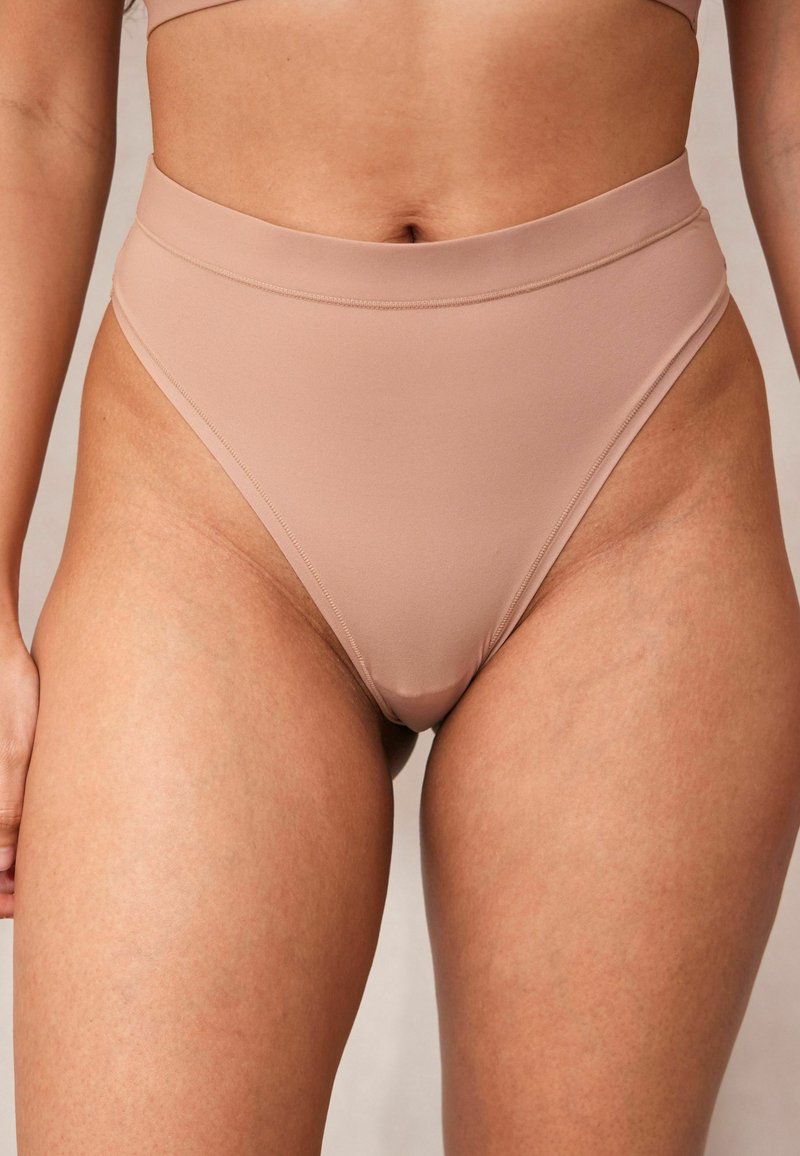 Bas de bikini taille haute en tissu nude. Texture lisse, avec un design sans coutures et des coutures minimales. Offre une ouverture de jambe haute.