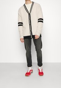 Osoba v krémovém kardiganu s černými pruhy, bílém tričku, tmavých rolled-up džínách a červených teniskách New Balance stojí na bílém pozadí.