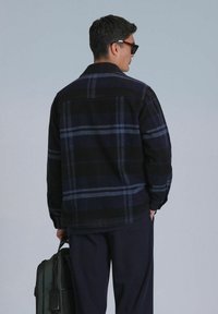Veste chemise à carreaux noir et marine avec col, manches longues et coupe décontractée. Portée avec un pantalon foncé et un sac vert.