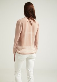 Blusa trasparente rosa chiaro a maniche lunghe con un motivo testurizzato, polsini arricciati e un orlo posteriore leggermente più lungo, indossata su pantaloni bianchi.