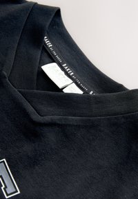 Sort bomuld t-shirt med V-hals design. Indvendig etiket viser "BAKER by Ted Baker" i hvid tekst med grønne accenter.