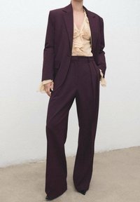 Costume bordeaux avec une veste cintrée et un pantalon à jambes larges, associé à un chemisier beige à volants. Tissu lisse, design structuré, silhouette élégante.