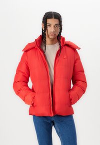Rød polstret jakke med høj krave, lynlås foran og to sidelommer. Har en glat overflade og horisontal quiltet design.