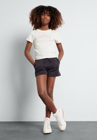 T-shirt crème à manches courtes avec le logo "ROXY" en rose, associé à un short noir. Le modèle porte des baskets montantes blanches avec des accents rouges.
