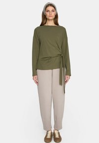 Oliwkowa bluzka z długim rękawem z węzłem, połączona z jasnobeżowymi spodniami i brązowymi butami na sznurówki. Gładki, elastyczny materiał, luźny krój.