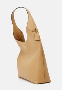 Sac à bandoulière en cuir fauve avec une large sangle et une petite étiquette attachée, coutures minimales, présenté en vue latérale inclinée sur fond blanc.