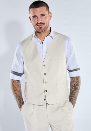 Man with short hair and tattoos on arms wearing a beige vest and pants over a white shirt with rolled sleeves, hands in pockets.