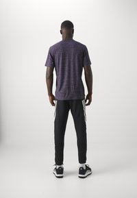 T-shirt sportivo viola con maniche corte, abbinato a pantaloni jogger neri con strisce bianche ai lati. La trama sembra liscia e leggera.