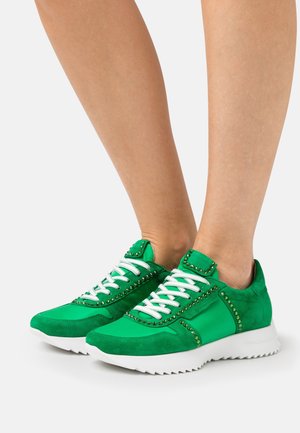 Piedi che indossano sneakers verde scuro in suede e tessuto, con lacci bianchi e decorazioni a borchie su suole bianche zigrinate.