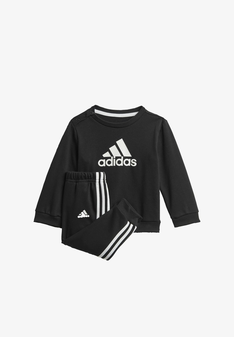 Čierna mikina Adidas s bielym logom, so zapínaním na pleciach; spárovaná s čiernymi nohavicami, s bielymi pruhmi pozdĺž strán.