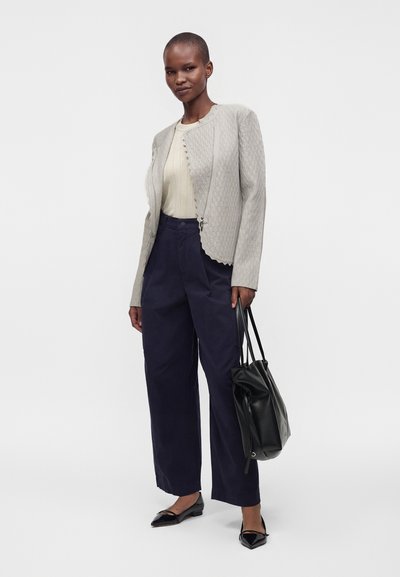 Femme debout, portant une veste légère texturée, un pull crème, un pantalon large bleu marine, des ballerines noires et un sac à main noir.