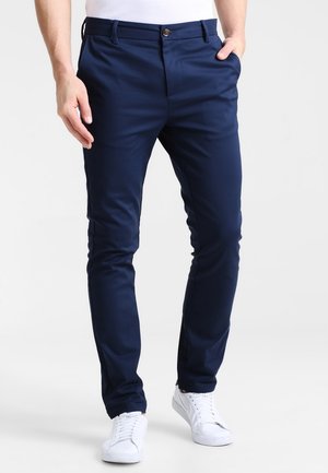Homme portant un pantalon slim bleu marine et des baskets blanches, debout avec une main dans la poche sur un fond uni.