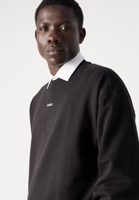 Felpa nera con collo a giro e logo "HUGO" bianco sul petto. Indossata sopra una camicia bianca con colletto, caratterizzata da una texture liscia.