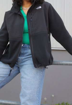 Osoba ubrana w czarną polarową kurtkę North Face na zielonym topie i jasnoniebieskich dżinsach, opierająca się o metalową balustradę na zewnątrz.