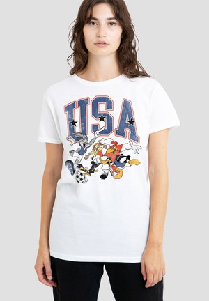 Junge Frau trägt ein weißes T-Shirt mit großem "USA" Text und Looney Tunes Figuren, die Fußball spielen, auf der Vorderseite.