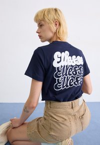 T-shirt bleu marine avec un grand logo blanc "Ellesse" dans le dos. Manches courtes et col rond. Associé à un short taille haute beige.