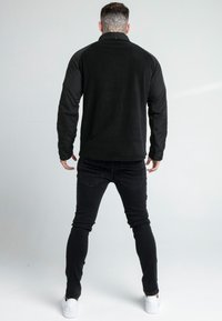 Giacca in pile nera con maniche in nylon nere a contrasto, colletto alto e vestibilità rilassata, abbinata a jeans scuri e sneakers bianche.