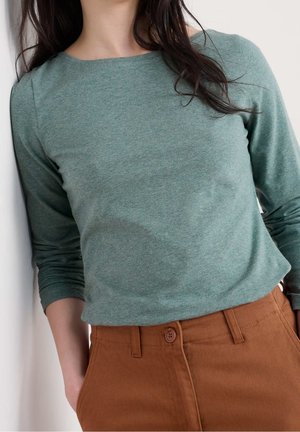 Kobieta w przygaszonym zielonym topie z długim rękawem i wysokim stanem rdzawych spodni z przodu z guzikami i kieszeniami, stojąca przy białej ścianie.