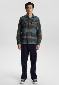 Camicia a bottoni con stampa camouflage e motivi geometrici blu, abbinata a pantaloni di velluto scosso scuri e sneakers nere.