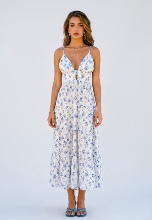Woman stands wearing a white sleeveless floral midi dress with thin straps and blue sandals against plain white background.