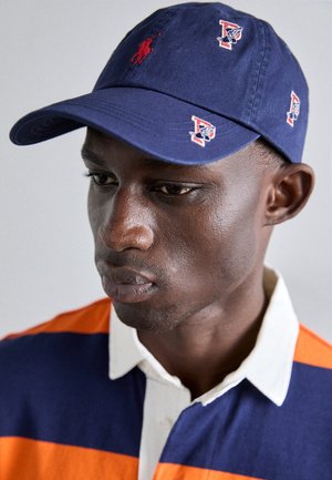 Jeune homme portant une casquette marine avec des logos rouges brodés et un polo rayé bleu et orange avec un col blanc.