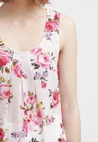 Top senza maniche floreale in tessuto leggero e traslucido. Presenta rose rosa e viola su uno sfondo crema con sottili pieghe sul collo.
