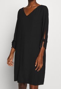 Czarna sukienka z dekoltem w kształcie litery V, z rękawami o długości trzy czwarte, z bocznymi rozcięciami zakończonymi wiązaniami. Gładki materiał, luźny krój, długość do kolan.