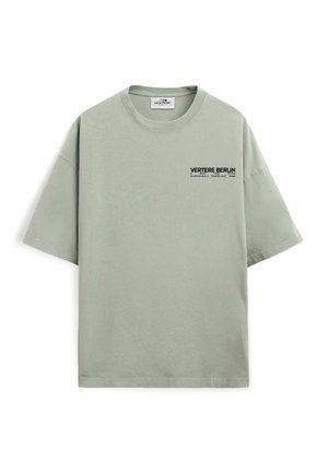 Junger Mann mit kurzen Haaren steht mit den Händen in den Taschen, trägt ein hellgraues oversized T-Shirt mit dem Schriftzug „Vertere Berlin“ und dunkle Hosen.