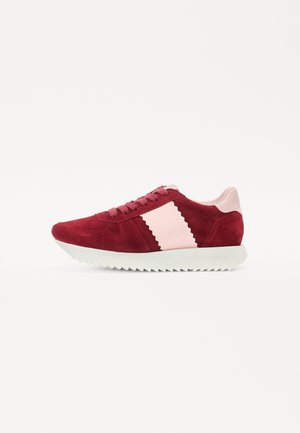 Sneaker en daim rouge avec des accents roses, présentant un design ondulé sur le côté, des lacets en tissu et une semelle blanche crantée.