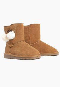Botas de tobillo de ante color beige con punta redondeada, que presentan un lazo decorativo y dos pompones cremosos y esponjosos en el lateral, suelas de goma y detalles de costura.