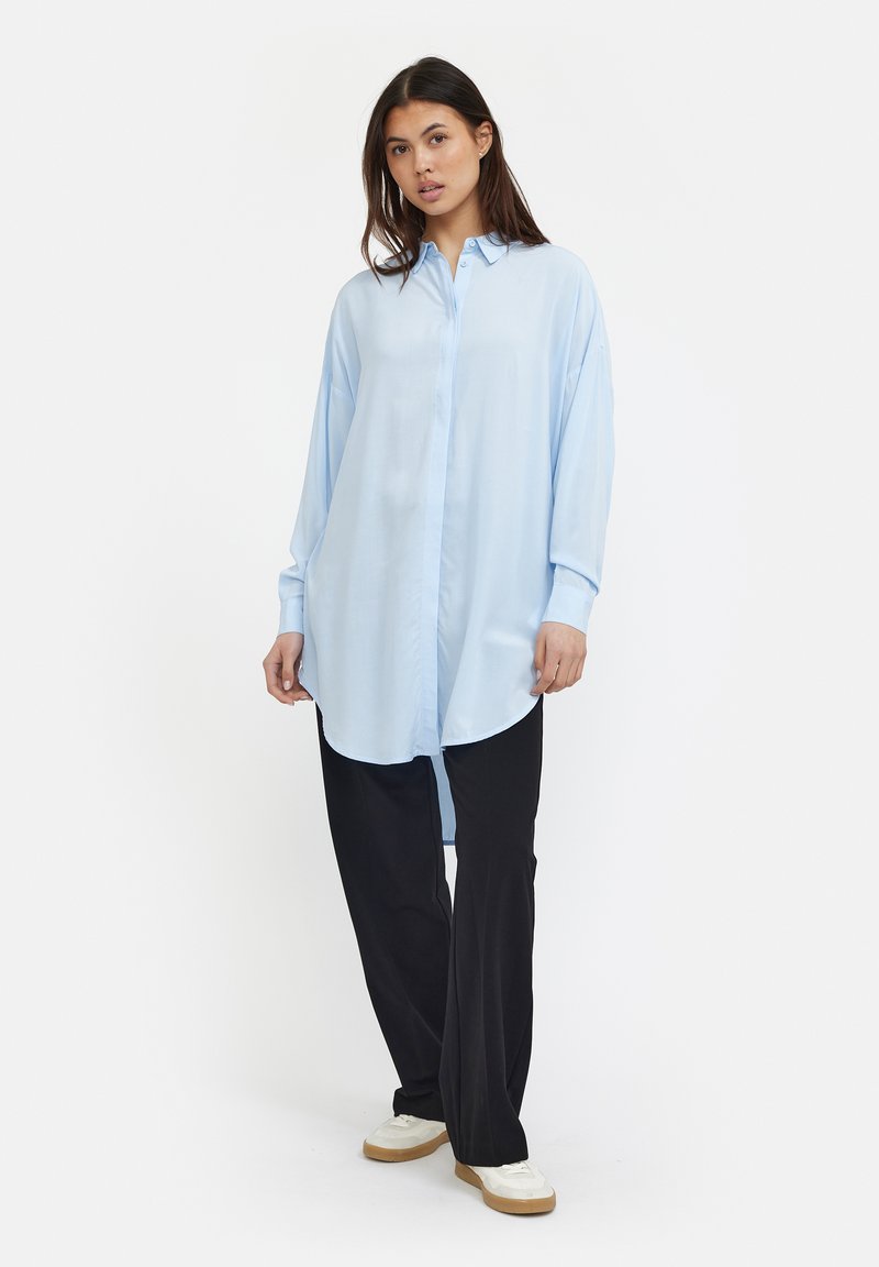 Camicia azzurra chiaro con maniche lunghe, bottoni e una vestibilità ampia, orlo arrotondato e spacchi laterali, abbinata a pantaloni neri a gamba larga e sneakers bianche.
