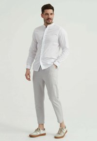 Chemise blanche à manches longues et col mandarin, associée à un pantalon slim gris clair et des baskets blanches avec des accents beiges.