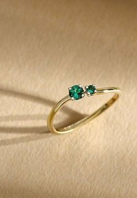 Guldring med två gröna ädelstensaccenter, en större än den andra, med en slät band och en polerad yta.