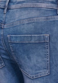 Vaqueros de denim azul con un lavado descolorido, que presentan dos bolsillos traseros contorneados con costuras beige. La tela tiene una textura suave.