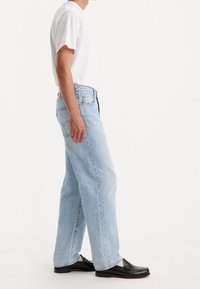 Person som bär ljusblå raka jeans, vit kortärmad skjorta och svarta lädersandaler, stående mot en neutral vit bakgrund.