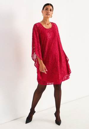 Röd spetsklänning med batwingärmar och rund halsringning, med detaljerade blommönster och en lös silhuett. Buren över svarta leggings och klackar.