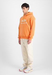 Sudadera con capucha naranja, con un bolsillo frontal y un logotipo blanco. Combinada con pantalones cargo beige claros y zapatillas deportivas multicolores.