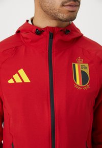 Detailný záber osoby v červenej zipsovej bunde so žltým logom Adidas a erbom Kráľovskej belgickej futbalovej asociácie na hrudi.
