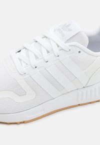  Zapatilla deportiva blanca con parte superior de malla, tres franjas características y una suela texturizada con un acento de goma en color marrón claro para mayor tracción.