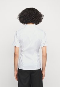 Chemise blanche à manches courtes avec col, coupe ajustée, texture lisse et poignets retroussés. Détails de coutures visibles à l'arrière.