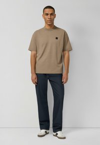 Brązowy bawełniany T-shirt z krótkimi rękawami, z małym czarnym logo. Noszony z ciemnoniebieskimi spodniami i białymi trampkami. Prosty fason i design.