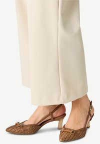 Décolleté intrecciata marrone con punta appuntita, corpo testurizzato, tacco piccolo e accessori metallici sul davanti. Abbinate a pantaloni color crema.