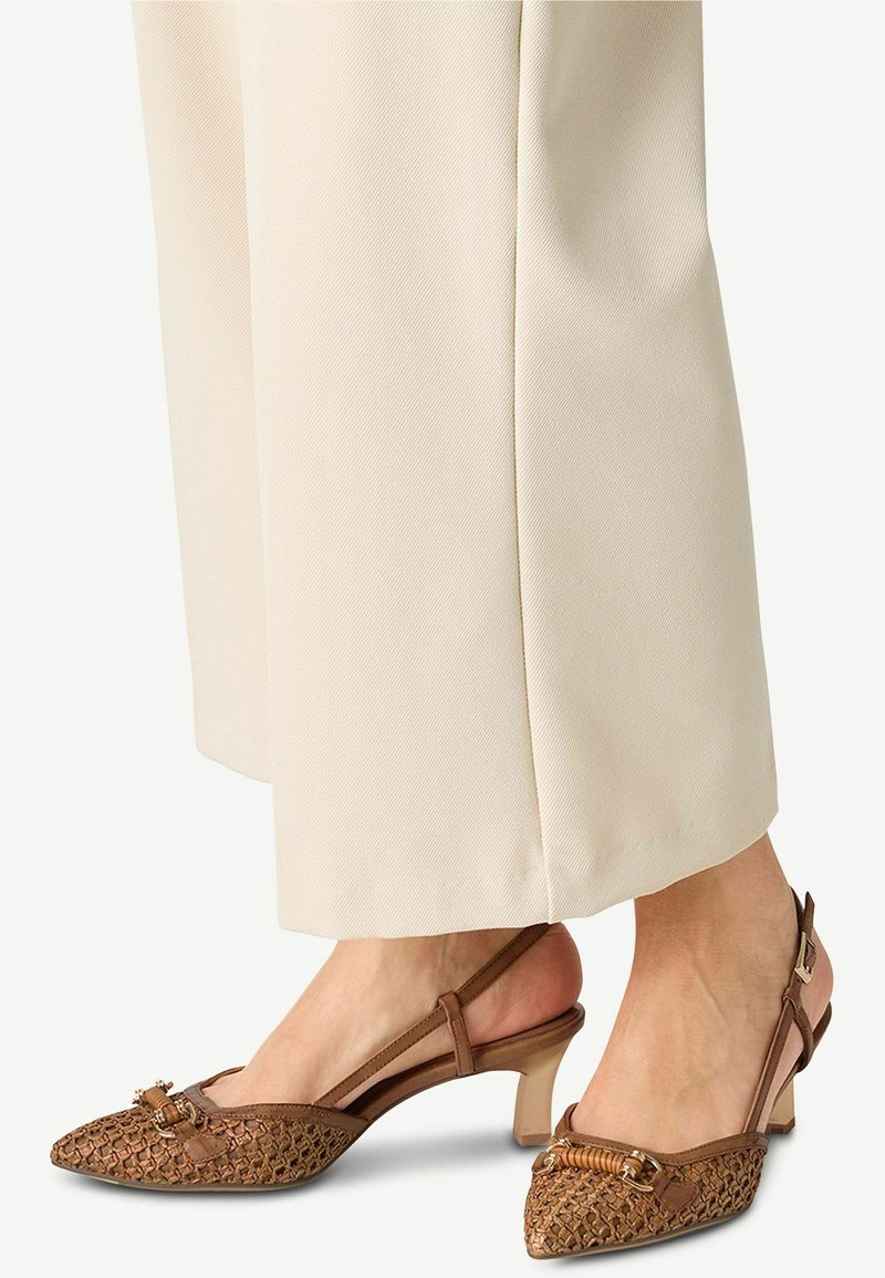 Décolleté intrecciata marrone con punta appuntita, corpo testurizzato, tacco piccolo e accessori metallici sul davanti. Abbinate a pantaloni color crema.