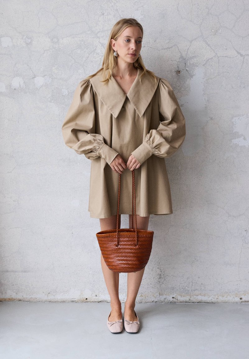 Γυναίκα που φοράει μπεζ oversized φόρεμα με μεγάλο γιακά και φουσκωτά μανίκια, κρατώντας μια καφέ πλεκτή τσάντα μπροστά σε έναν γκρίζο τοίχο.