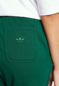 Zelené tepláky s zadní kapsou a tonálním vyšitým logem. Látka má hladkou texturu a elastický pas.