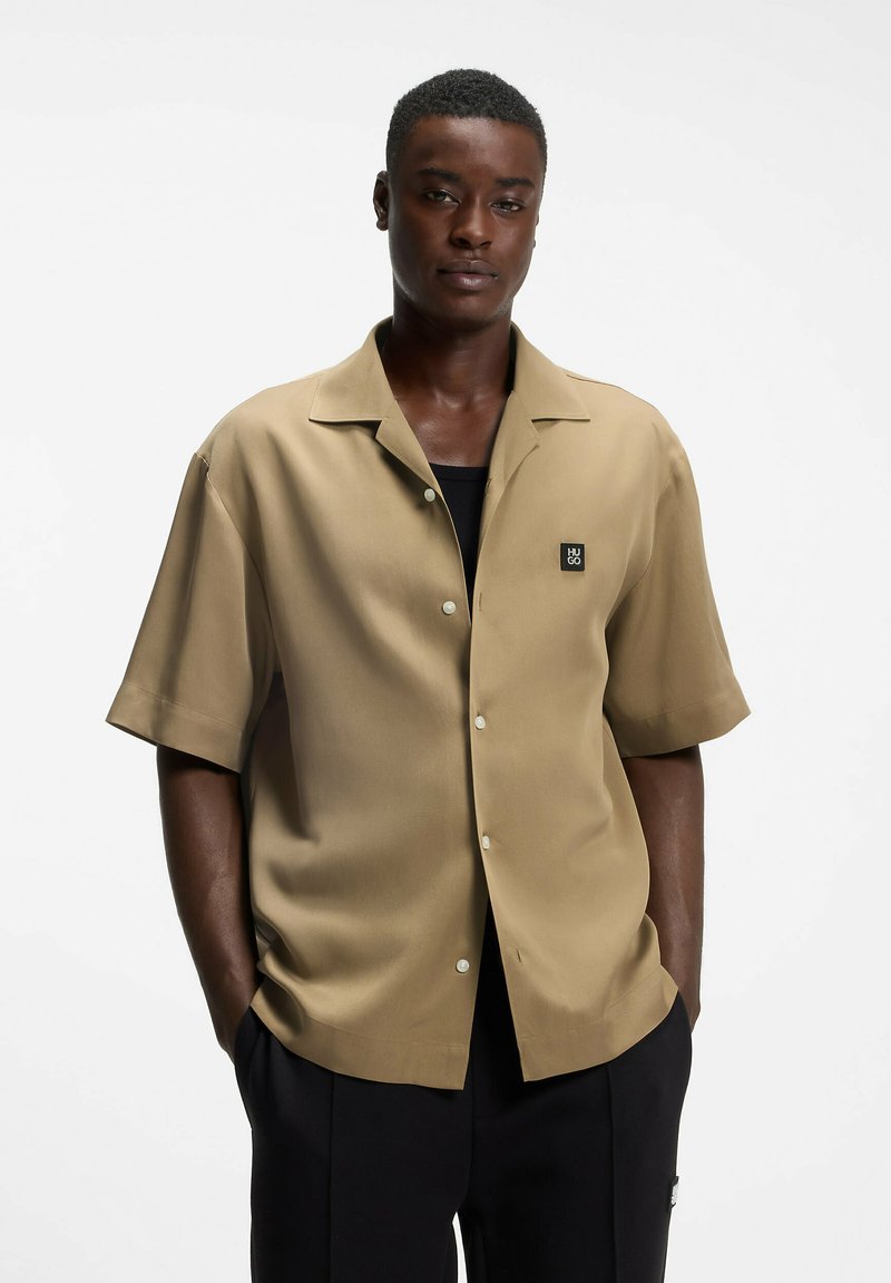 Homme debout, les mains dans les poches, portant une chemise beige à manches courtes boutonnée sur un haut noir et un pantalon noir sur fond blanc.