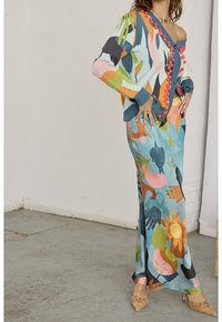 Kolorowy sweter z dzianiny z akcentami w postaci pomponów, zestawiony z wzorzystą sukienką maxi z motywem rąk i kwiatów w wielu kolorach.