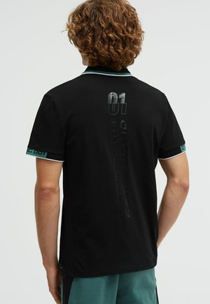 Person mit lockigem Haar trägt schwarzen Poloshirt mit vertikalem Text und Nummer "01" auf der Rückseite, mintgrüner Besatz an Ärmeln und Kragen.