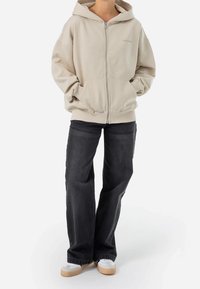 Beige zip-up hoodie med huva, kängurufickor och ribbade manschetter. Matchad med mörkgrå vida jeans och vita skor.