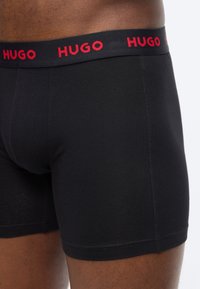 Μαύρα αντρικά σλιπ boxers που φοριούνται από έναν άνθρωπο, με μαύρη ζώνη μέσης που έχει επαναλαμβανόμενο κόκκινο λογότυπο "HUGO".